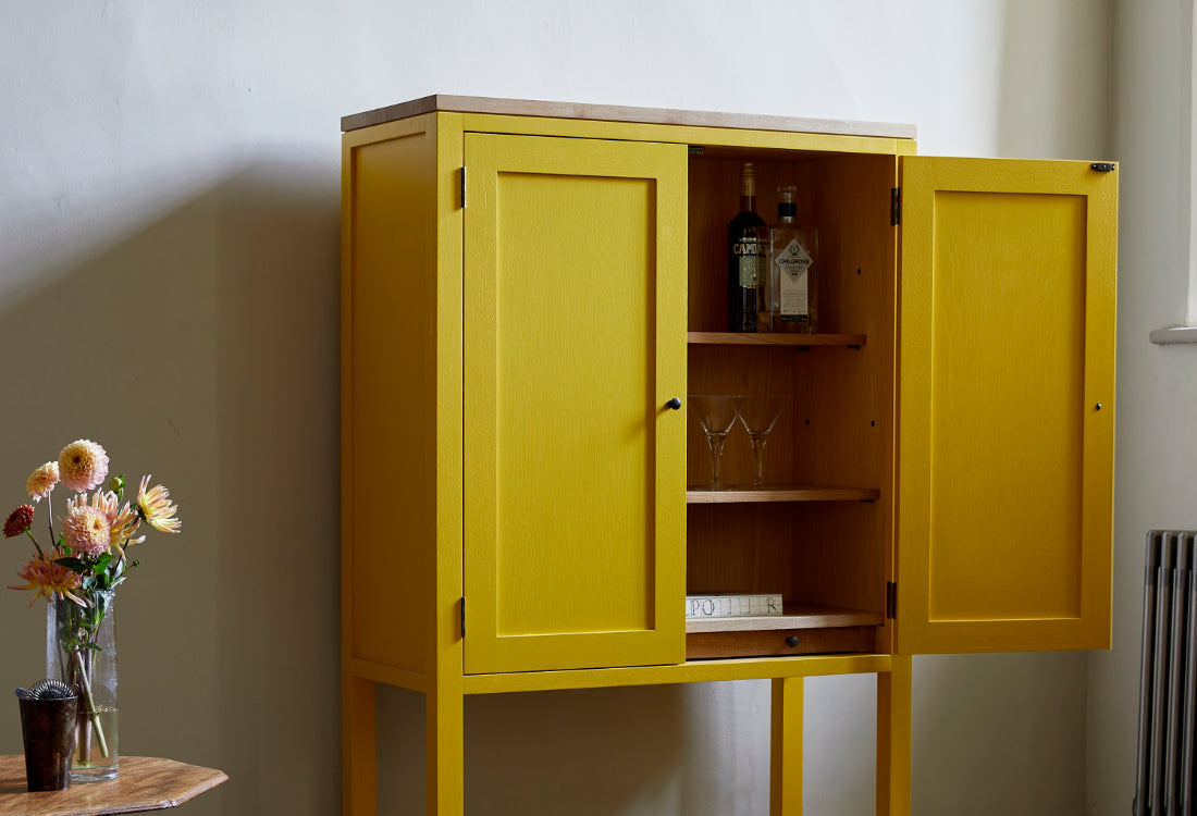 Yellow painted shaker style cupboard door open showing shelves with alcohol drinks bottles and cocktail glasses in room with large window traditional radiator and vase with flowers.