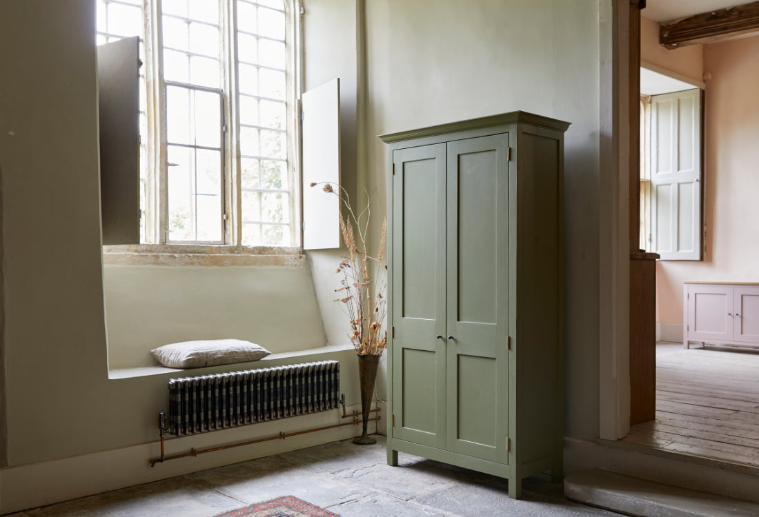 Tall freestanding two door shaker style green cupboard open with shelves of books and linens large leaded windows with shutters and pink painted sideboard in background.
