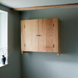 Oak cabinet with two doors, and metal knobs. Shot in room against blue wall, Vase with dried flower in vase on windowsill.