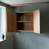 Oak cabinet with two doors, and metal knobs. Picture in room against blue wall, Vase with dried flower in vase on windowsill. Both doors open showing woven wood back. Three glass vases on shelf in cabinet.