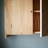 Close up of Oak cabinet with two doors, one open, and metal knobs. Pictured against blue wall. Open door reveals woven wood back,