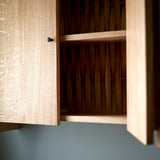 Close up of Oak cabinet with two doors, one open, and metal knobs. Pictured against blue wall. Open door reveals woven wood back,