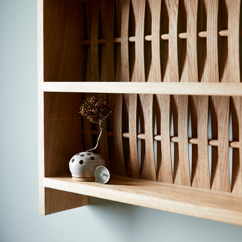 Close up of shelve unit in oak wood. Three shelves. Woven wood back. Pictured on wall in blue room. Two small ceramic dishes on shelf.