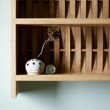Close up of shelve unit in oak wood. Three shelves. Woven wood back. Pictured on wall in blue room. Small ceramic dishes and vintage compass on shelf.