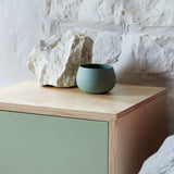 Close up of single door ply wood contemporary bedside cabinet with green painted door. Against painted white stone wall with large rustic piece of natural chalk and ceramic bowl on cabinet.