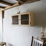 Two door ply wood contemporary wall cupboard with reeded glass doors and black metal door knobs. With simple white ceramic vase and jug on top with traditional wooden chair and dried pampas grass stems in foreground.