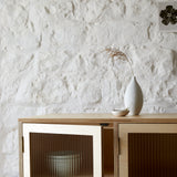 Close up of cabinet corner showing reeded glass door. Simple white ceramic vase and bowl on top against a white painted stone wall.