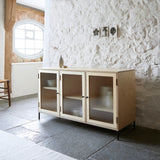 Ply wood sideboard with three reeded glass doors on angle against a white painted stone wall and on black flag stone floor.
