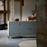 Three door ply wood contemporary side board with blue grey painted doors, black metal knobs and feet shown in farmhouse room setting.