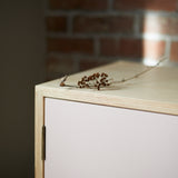 Angle corner close up shot showing ply wood joint detail, pink painted door, hinge and dried seed heads on top. Blurred exposed brick wall in background.