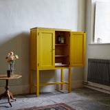 Yellow painted two door shaker style cabinet on tall legs with oak work top. Door open revealing shelves with drinks bottles and glassed in room with flagstone floor traditional radiator large sash window patterned rug pedestal table with vase of flowers.