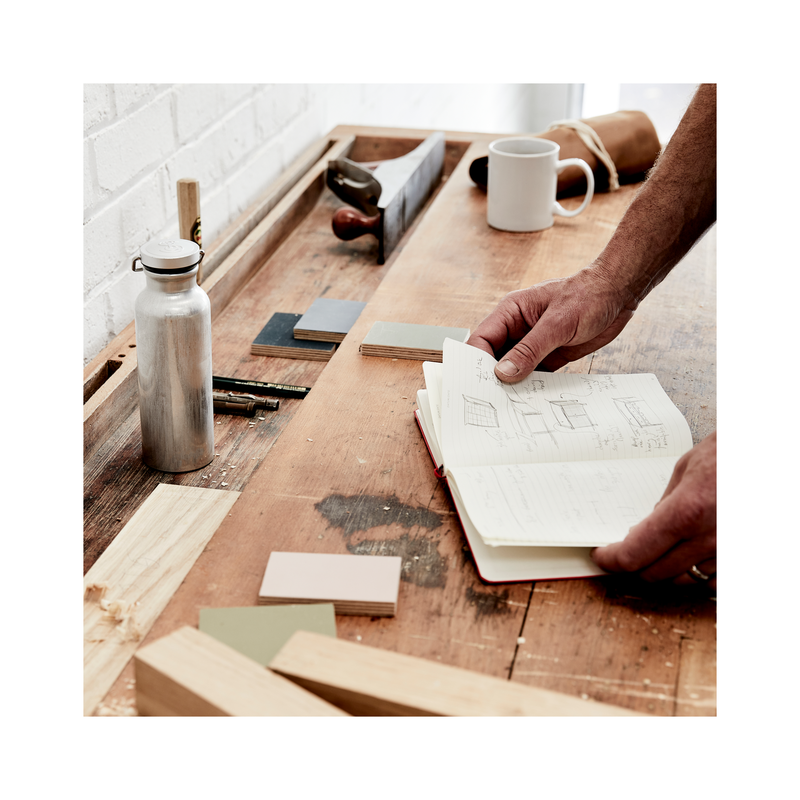 Work bench with wood off cuts, metal drinks bottle, white mug. Note book held open with two hands.