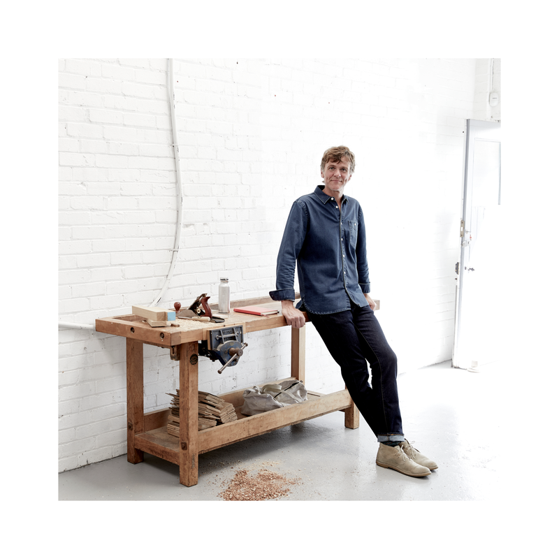 Tall man in jeans and blue shirt leaning against a traditional carpenters workbench with tools on. Grey painted concrete floor and white painted brick walls.