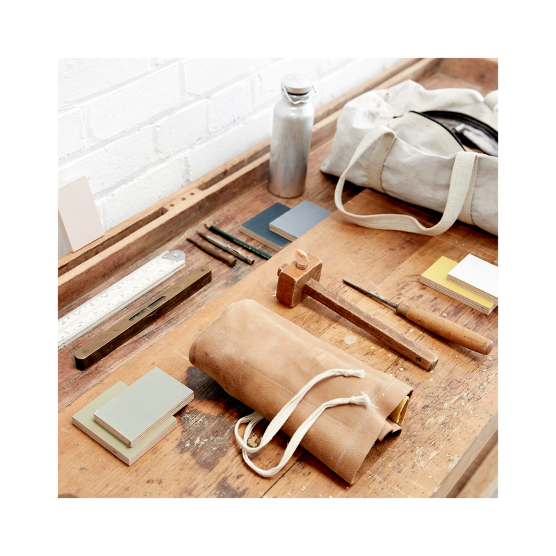 Work bench with blue canvas tool bag, leather tool roll, colour samples, mortice scribe, chisel and metal water bottle.