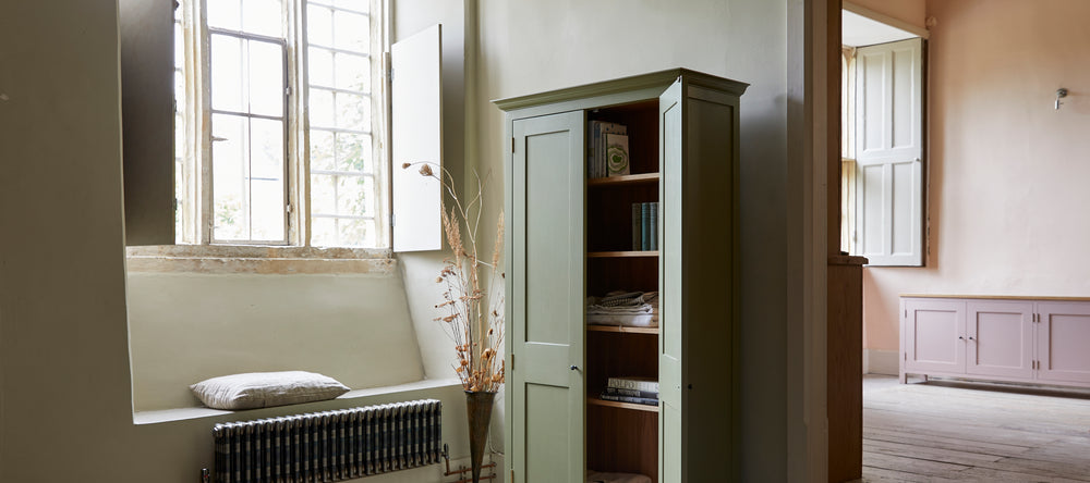Tall freestanding two door shaker style green cupboard open with shelves of books and linens large leaded windows with shutters and pink painted sideboard in background.