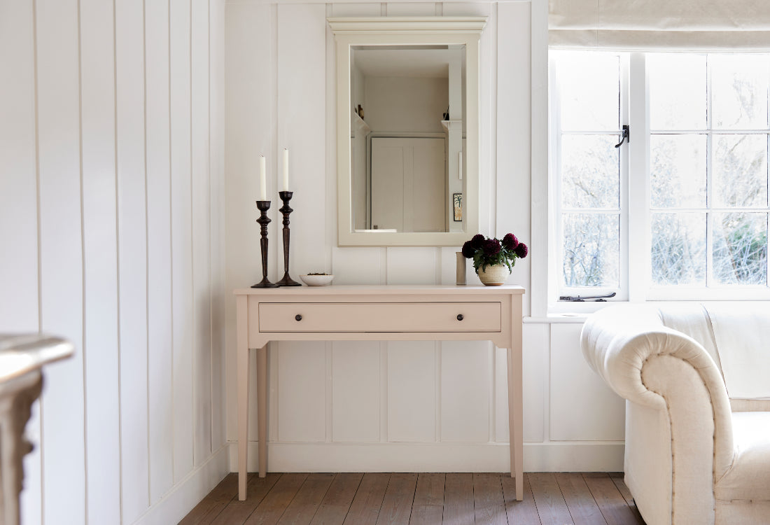 Light wooden painted console table with mirror and candles in a bright room with a window.