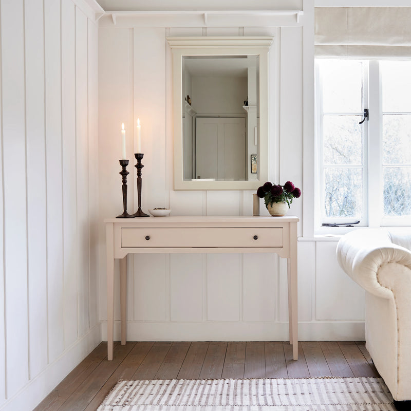 A light pink console table with a single drawer and small dark knobs, styled in a bright white room with a mirror, candles, flowers, and a window.