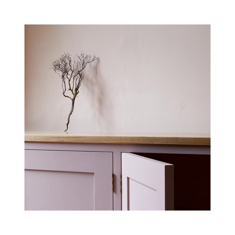 Close up of pink painted sideboard with oak worktop dried flower stems on top leaning agains soft plaster coloured wall.