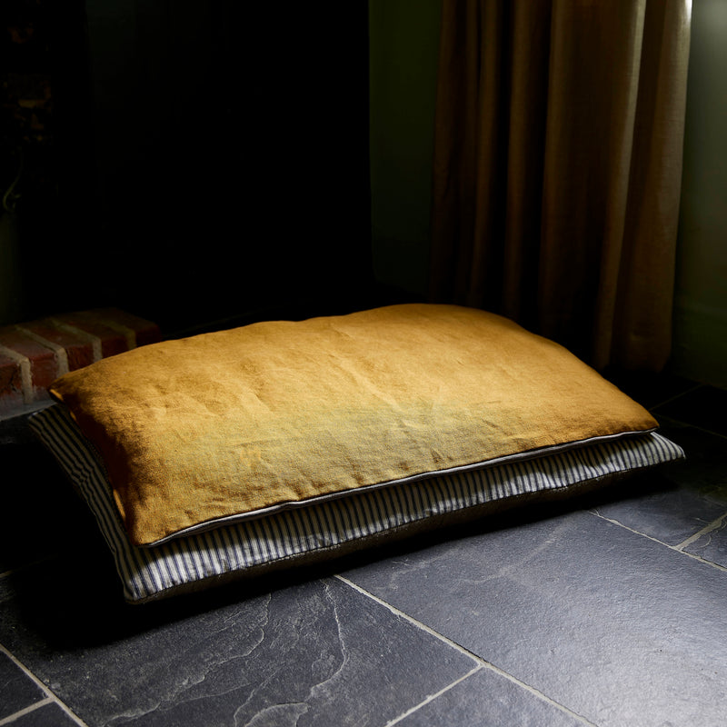 Dog bed ochre linen topper on cotton ticking stripe floor cushion. In room with black slate floor and linen curtains
