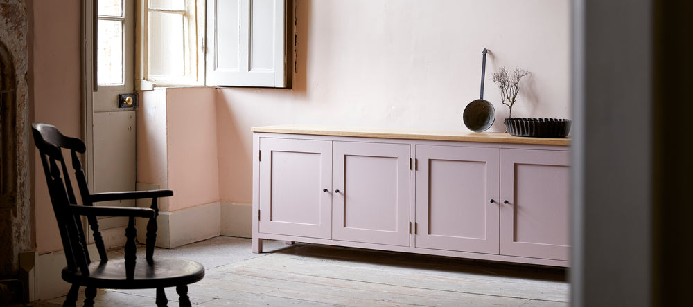 Four door pink painted sideboard with oak top in room with stripped wood floor boards large sash windows and traditional wooden captains chair.