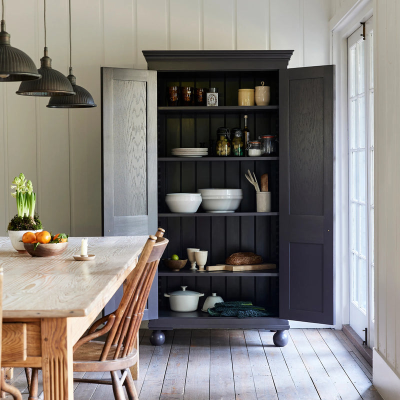 A tall brown wooden pantry cabinet with open doors, styled in a bright kitchen with hanging pendant lights, a wooden dining table, and white panelled walls.