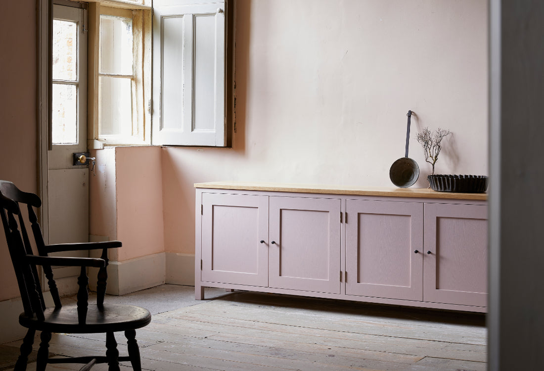Four door pink painted sideboard with oak top in room with stripped wood floor boards large sash windows and traditional wooden captains chair.