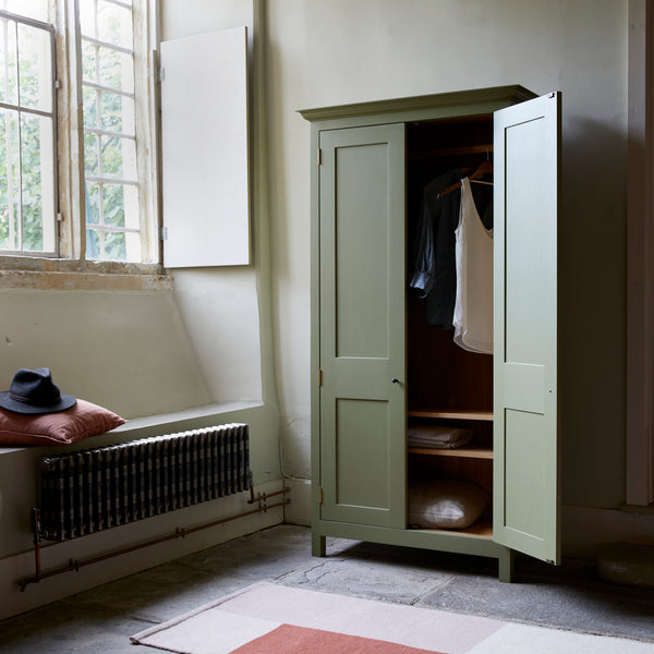 Green painted Armoire shaker style cupboard in room setting with one door open showing clothes on rail and on two shelves. Flagstone floor and large leaded window with shutters. Traditional radiator and window seat with cushion and hat
