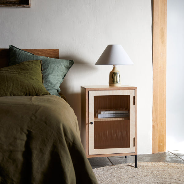 Ply wood bedside cabinet with glazed door with books on internal shelf. Pottery lamp with white fabric shade on top of cabinet. On a jute rug with bed dressed in green linen.