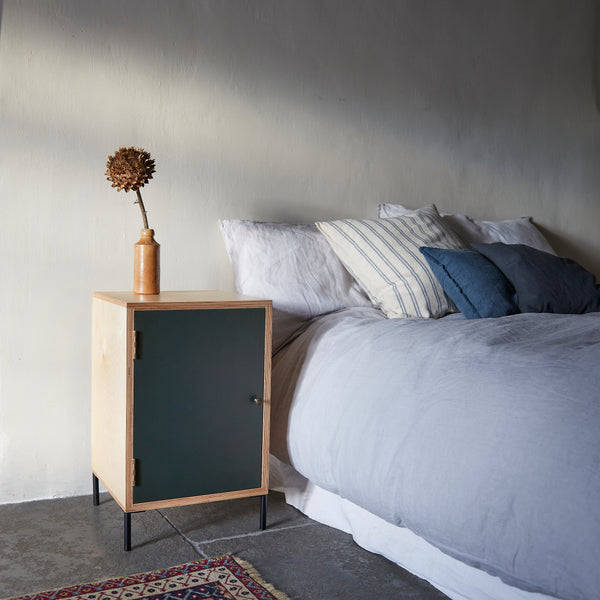 Ply wood bedside cabinet with dark grey painted door, black metal door knob and black metal feet with stoneware vase with large dry flower head in on top,. Next to bed with blue linen bedding and ticking pillows on black slate floor with patterned rug.