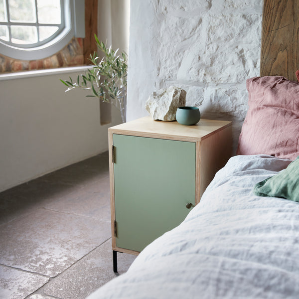 Single door ply wood contemporary bedside cabinet with green painted door, black metal door knobs and black metal legs. In room with flagstone floors, linen covered bed and sash window. Large rustic pieces of natural chalk and ceramic bowl on cabinet.