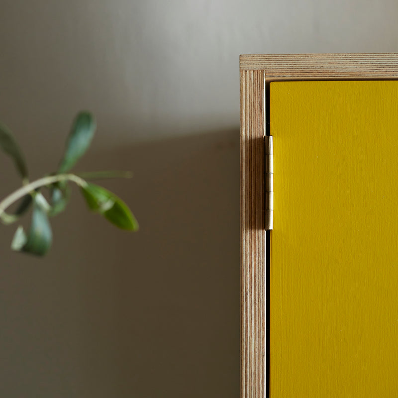 Close up of corner of ply wood cabinet showing joint detail and bright yellow painted door and hinge.