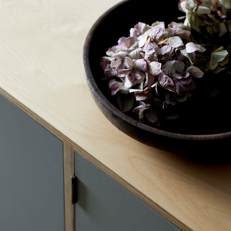 Dark wooden bowl with dried hydrangea flower heads on top of birch ply wood cabinet top with two painted blue grey doors.