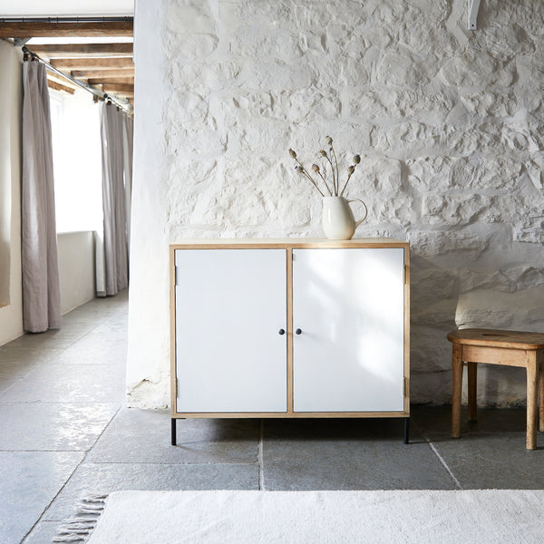 Two door contemporary ply wood sideboard. Doors painted white with black door knobs in farmhouse room setting against a white painted stone wall and on black flagstone floor with a white rug in foreground. Simple wooden stool to side and windows with floor length grey curtains.