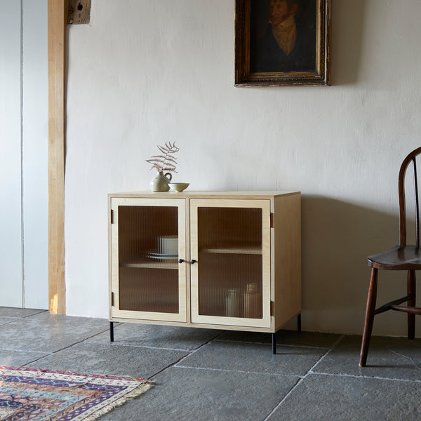 Contemporary glazed ply wood cabinet with reeded glass and black slim metal legs. In room with black flagstone floor, old Windsor chair, oil painting of portrait on wall and patterned rug in foreground.