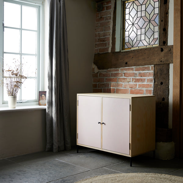 Two door ply wood contemporary side board with pink painted doors, black metal knobs and feet. Shown in a Georgina room by sash casement window with light flooding in and against an expose brick and beam wall with stained glass above.