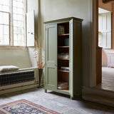 Tall freestanding wooden Shaker style two door cupboard with cornice and wooden legs, painted apple green. One door open revealing four shelves and oak interior. One door open revealing books and linens on shelves. Shown in a room with black flagstone floor large sash windows patterned rug on floor.