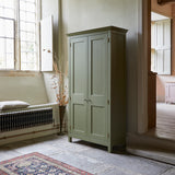 Tall freestanding wooden Shaker style two door cupboard with cornice and wooden legs, painted apple green. Shown in room with black flagstone floor and large sash windows with shutters. Patterned rug in foreground.