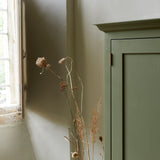 Corner of green painted tall freestanding cupboard, showing cornice and brass hinge. Dried flower seed heads to side.