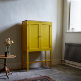 Yellow painted two door shaker style cabinet on tall legs with oak work top in room with flag stone floor traditional radiator large window and pedestal table with vase of flowers.