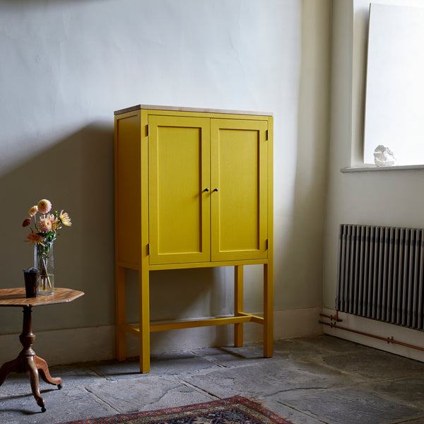 Yellow painted two door shaker style cabinet on tall legs with oak work top in room with flag stone floor traditional radiator large window and pedestal table with vase of flowers.