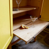 Yellow painted cabinet doors open revealing oak interior and sliding shelf with lemon wedges and cocktail glasses.