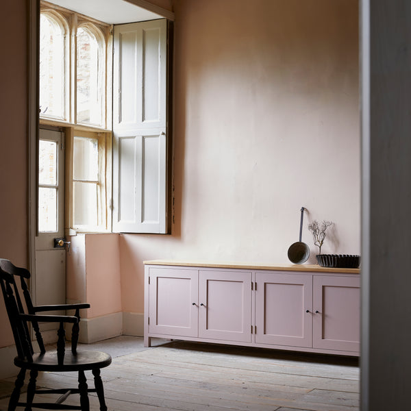 Pink painted four door shaker style sideboard with black metal door knobs and oak worktop. In room with stripped floor boards, large sash windows with shutters traditional wood captains chair and vintage large metal tart tin on top.