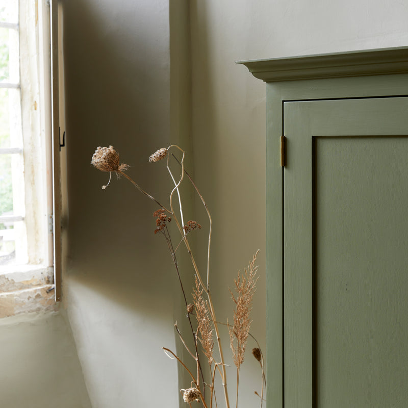 Close up of green painted shaker style cupboard showing cornice and brass hinges. Leaded window and dried seed heads.