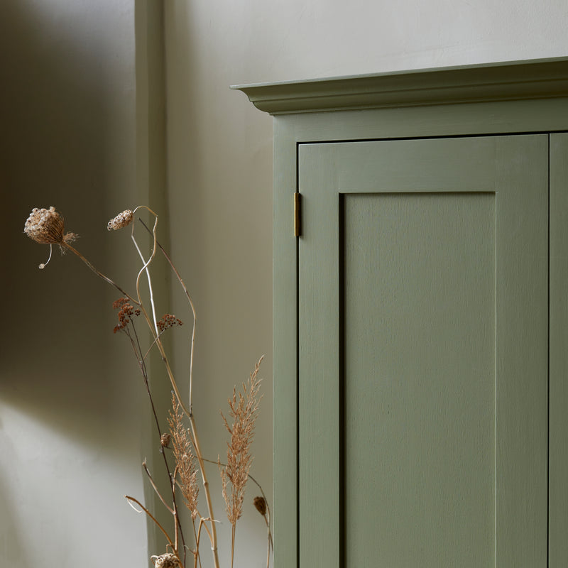 Close up of green painted shaker style cupboard showing cornice and brass hinges. Dried seed heads in foreground.