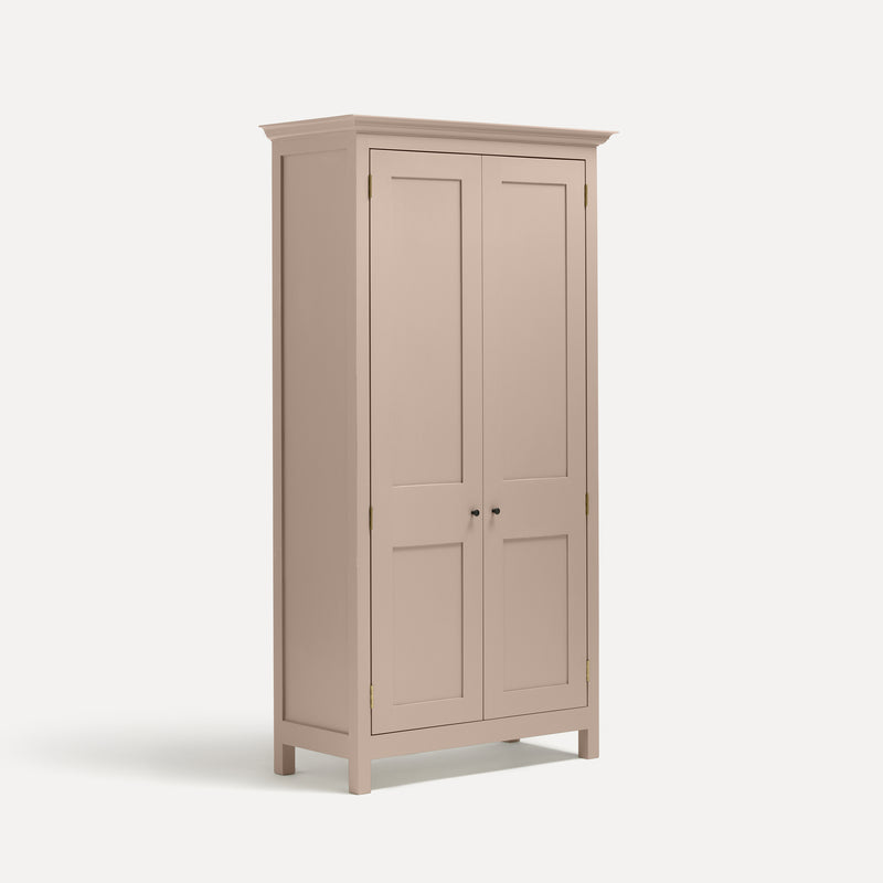 Pink painted freestanding tall cupboard Shaker style with panelled doors black metal knobs. Shown at an angle.