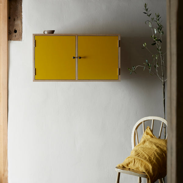 Wall cupboard with two bold yellow painted doors and black metal door knobs on wall with olive tree to side and traditional Windsor chair with yellow linen cushion.